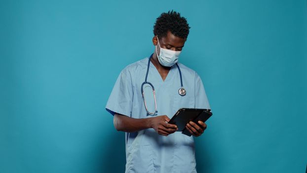 Medical assistant using digital tablet while wearing face mask and stethoscope in studio. Nurse with protection against coronavirus pandemic and blue uniform, looking at device screen