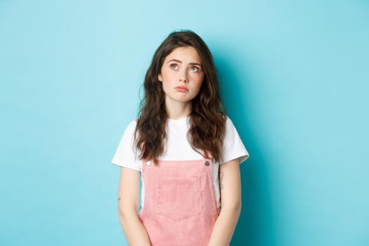 Portrait of sad and gloomy young brunette girl with glamour make up, looking at upper left corner upset and thoughtful, feeling moody, standing against blue background.