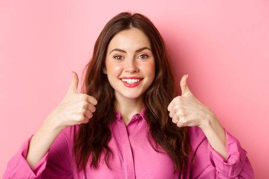 Close up of cheerful young lady fully approve your choice, praising something good, showing thumbs up and smiling pleased, pink background.