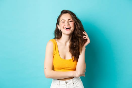 Summer and lifestyle concept. Portrait of attractive woman laughing and playing with her hair, standing in yellow cropped top, going on holiday, standing against blue background.