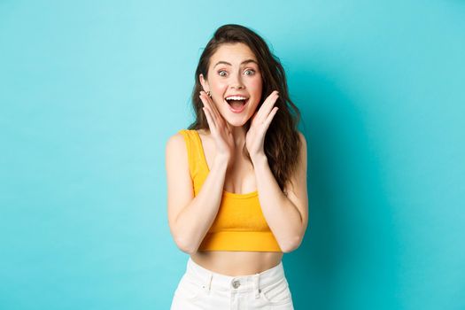 Summer and lifestyle concept. Young cheerful woman look with surprised face at camera, react to awesome good news, smiling and cheering, standing against blue background.