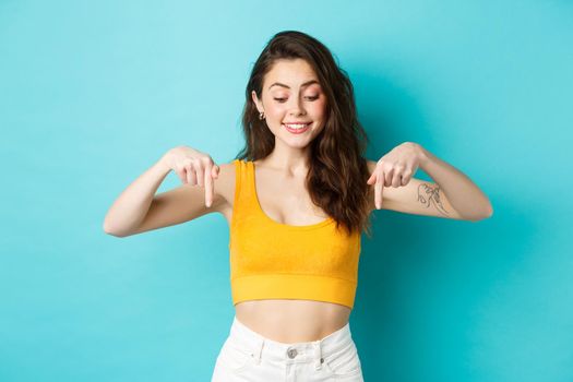 Portrait of stylish natural girl in summer crop top, pointing and looking down with dreamy smile, seeing something interesting on bottom advertisement, blue background.