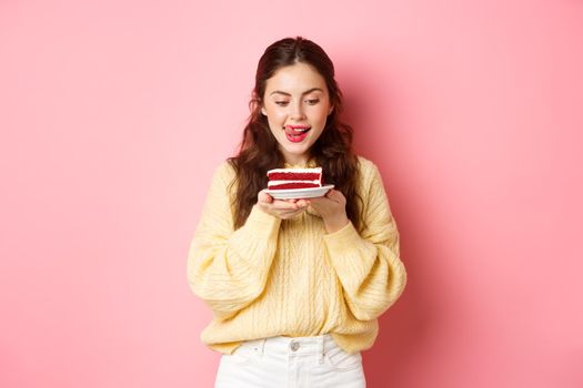 Young slim girl lick her lips from temptation, looking at delicious piece of cake with yearning to bite it, standing with dessert against pink background.