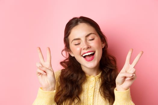 Positive emotions. Beautiful young woman showing peace v-sign and smiling carefree, posing with eyes closed and happy face, standing against pink background.