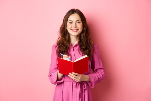 Cheerful girl smiling and writing down notes, holding her planner, plan schedule or grocery list, standing with diary against pink background.