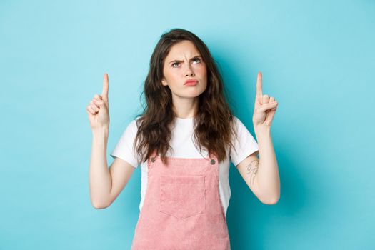 Grumpy cute brunette girl frowning displeased, sulking and looking disappointed up, pointing fingers on top, standing against blue background.