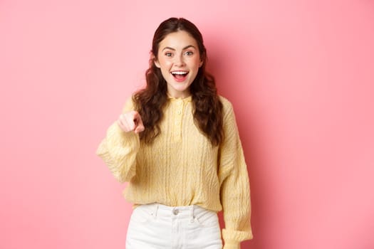 Young brunette woman laughing, smiling amazed and pointing at camera, choosing you, standing against pink background. Copy space