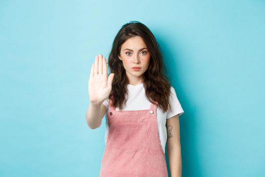 Young brunette woman with curly hairstyle, raising hand in block gesture, say stop or no, refuse bad offer, rejecting something, standing against blue background.
