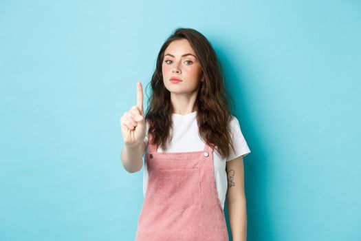 Rule number one. Serious young beautiful girl shaking finger in disapproval, scolding bad decision, make block or taboo gesture, standing against blue background.