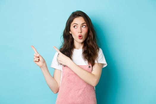 Glamour summer girl pucker lips and say wow, pointing and looking left at interesting promo deal, showing an advertisement, standing against blue background.