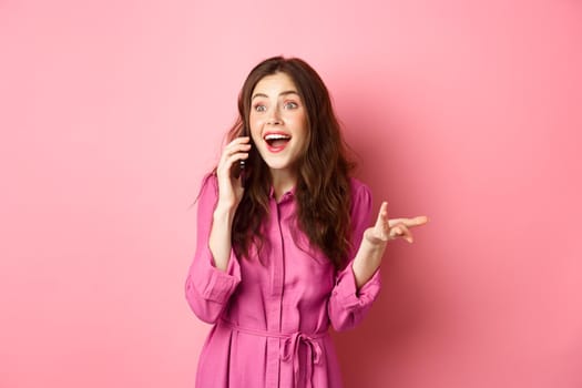 Cheerful brunette woman talking on mobile phone, gesturing and laughing, having casual funny conversation, standing over pink background.