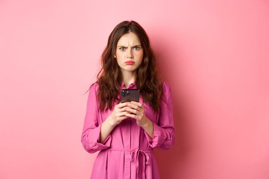 Angry woman frowning and looking confused after reading mobile phone news, standing puzzled and upset against pink background. Copy space