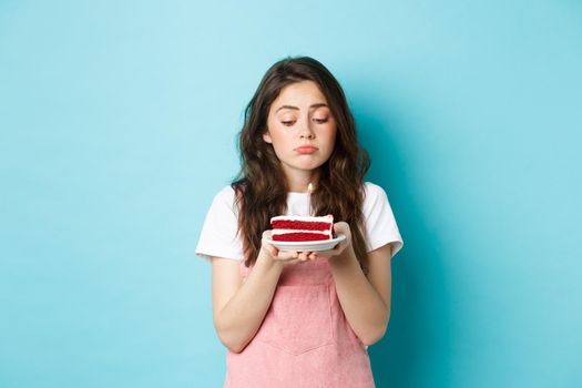 Holidays and celebration. Sad cute girl celebrating her birthday alone, holding cake on plate with one candle and sulking upset, standing lonely against blue background.