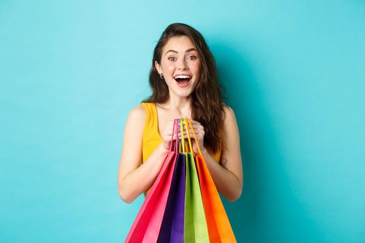 Portrait of surprised young woman look amazed at camera, holding shopping bags, see discounts in store, standing over blue background.