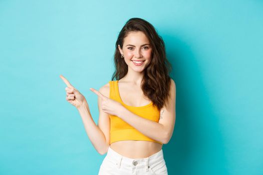 Portrait of stylish girl in colorful summer outfit, pointing fingers left at promo offer, showing your logo, smiling happy at camera, standing over blue background.