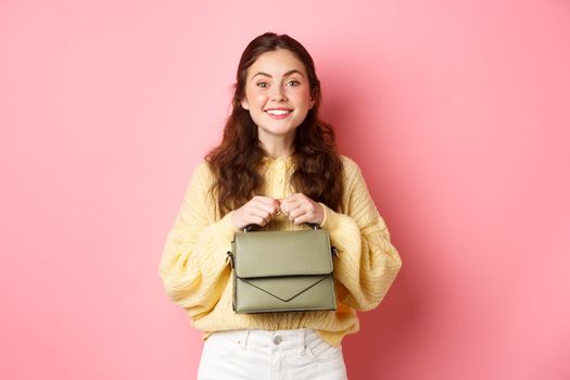 Beautiful lady holding her purse in hands near chest, smiling and looking excited at camera, waiting for something with temptation, standing against pink background.