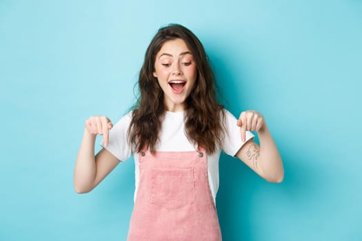 Happy beautiful girl in summer overalls, pointing fingers down, looking excited at item banner, checking out advertisement, standing over blue background.