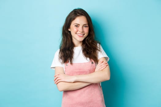 Happy smiling girl with cute blush, wearing summer clothes and beaming from joy, holding arms crossed over chest, standing against blue background.