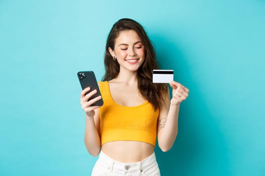 Stylish girl with plastic credit card smiling, paying for online order, shopping in smartphone, standing against blue background. Copy space