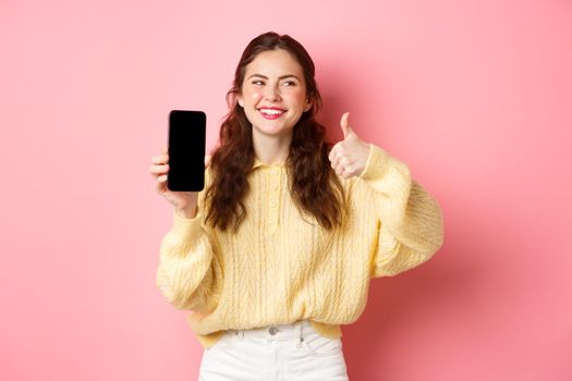 Image of beautiful woman laughing and looking aside, while rating an app, showing thumbs up and empty phone screen, recommending online promo, standing against pink background.