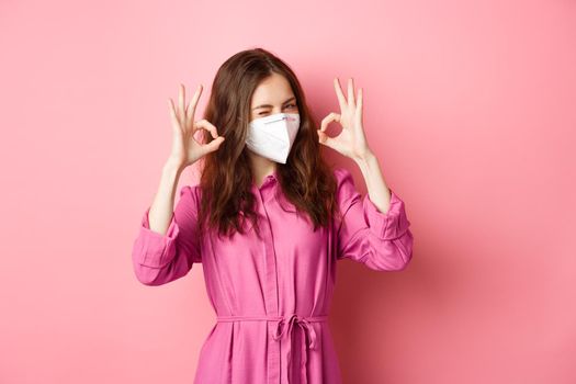 Covid-19, pandemic and lifestyle concept. Cheerful girl shows okay sign, wears medical respirator as preventive measure from corona, pink background. Copy space