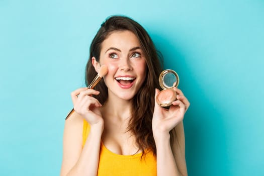 Beauty. Close up of beautiful young woman look upper left corner and smile while applying blushes on cheeks with make up brush, standing happy against blue background.