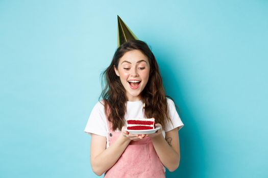 Holidays and celebration. Excited woman celebrating birthday, blowing candle on cake, wearing party cake and having fun, standing over blue background.