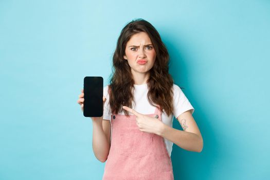 Skeptical and upset young woman frowning, sulking disappointed while pointing at empty smartphone screen with bad online offer or app, standing against blue background.