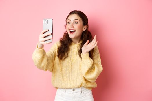 Image of woman video chatting with surprised face, receive amazing news, gasping excited, seeing something in smartphone screen, standing against pink background.