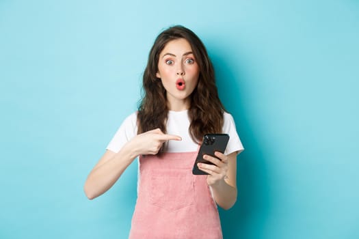 Portrait of excited woman pointing finger at smartphone, telling about online promo offer, showing advertisement on phone, standing against blue background.