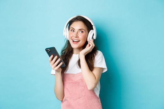 Image of young woman put on headphones and pick music on smartphone, holding phone, listening song, standing over blue background.