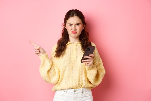 Displeased girl grimacing, frowning upset and pointing finger aside at left copyspace while holding smartphone, standing over pink background.
