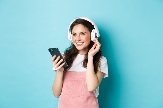 Stylish brunette girl in headphones holding smartphone, listening music, picking track in streaming app, standing over blue background.