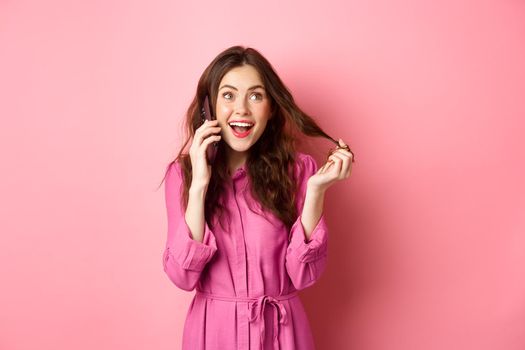 Glamour talkative girl playing with hair, making order, calling someone on phone, talking on smartphone, standing against pink background.