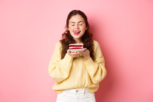 Woman holding delicious cake, licks her lips with closed eyes and dreamy face, wants to eat tasty dessert, standing against pink background.