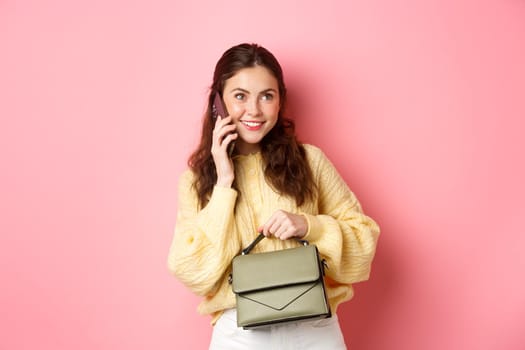 Young glamour girl holding her purse, talking on smartphone and smiling, having casual conversation on phone, calling someone, standing over pink background.