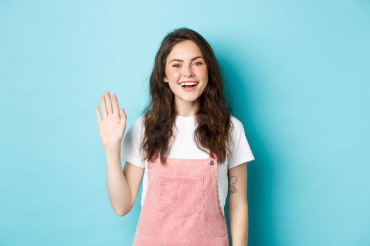 Hello nice to meet you. Beautiful friendly girl waving hand to say hi, looking happy and smiling while greeting you, giving warm welcome, standing over blue background.