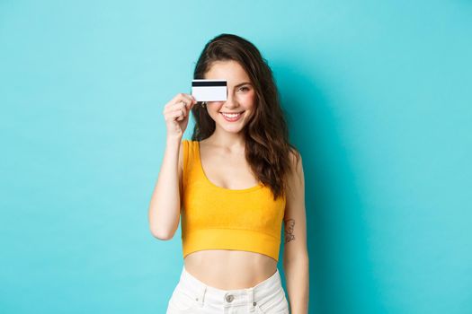 Young stylish woman showing plastic credit card over eye, looking pleased adn confident, smiling at camera, standing over blue background.