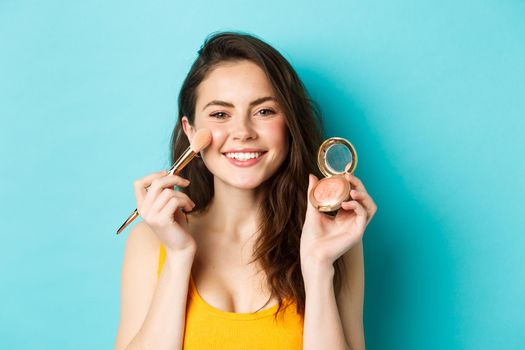 Beauty. Young attractive woman smiling, applying make up with brush, showing blushes at camera, standing over blue background.