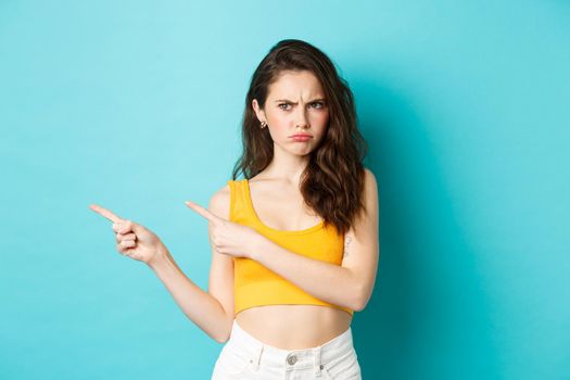Summer lifestyle concept. Angry and moody young woman looks jealous, pouting about something unfair, pointing and staring left at copy space, standing against blue background.
