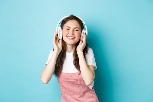 Cute caucasian girl in spring outfit, listening music in headphones, smiling pleased at camera, standing over blue background.