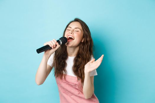 Carefree beautiful woman perform song, singing in microphone with passion, playing karaoke, standing over blue background.