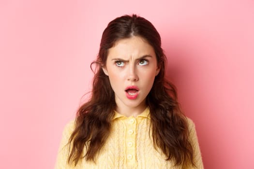 Young brunette woman with confused face, trying figure something out, frowning and looking up while thinking, standing pensive against pink background.