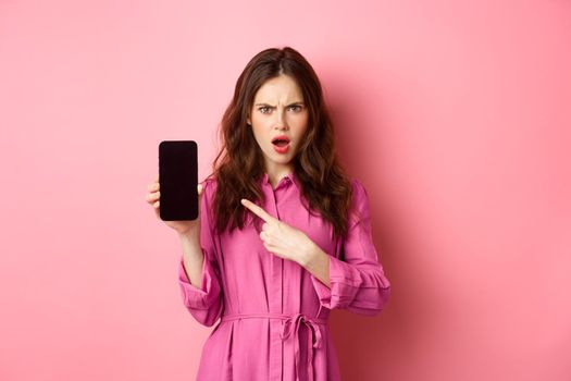 Technology concept. Angry girl points at her smartphone screen and staring judgemental at camera, demand answers or explanation, standing over pink background.
