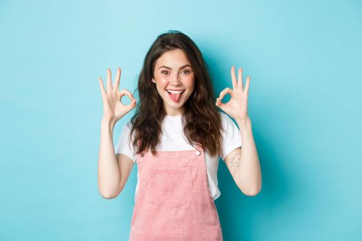 Portrait of excited brunette girl with curly hairstyle, showing okay signs, praise perfect choice, recommend good thing, give OK sign, standing over blue background.