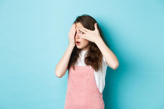 Portrait of shocked brunette girl gasping startled, covering eyes with hands but peeking through fingers at something embarrassing, standing against blue background.