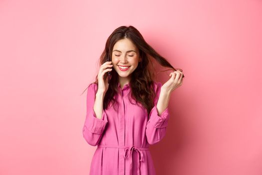 Beautiful young woman calling someone, laughing and smiling during phone call, talking to friend and playing with hair strands, standing against pink background.