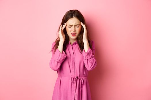 Woman feeling pain in head, touching temples and frowning, suffering migraine, being dizzy, standing with headache against pink background.