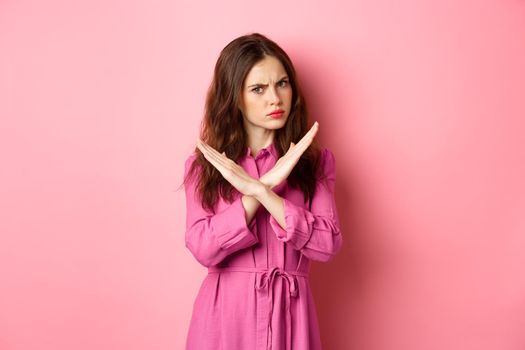 Displeased angry woman blocking offer, showing cross stop gesture, saying no and shaking head in negative reply, standing over pink background.