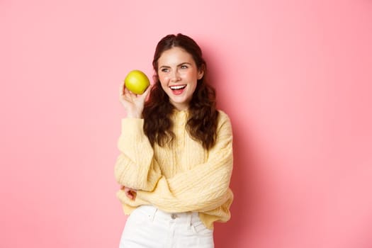 Healthcare and lifestyle concept. Cheerful smiling woman, showing her white perfect smile and green apple near face, eating healthy fruits, standing against pink background. Copy space